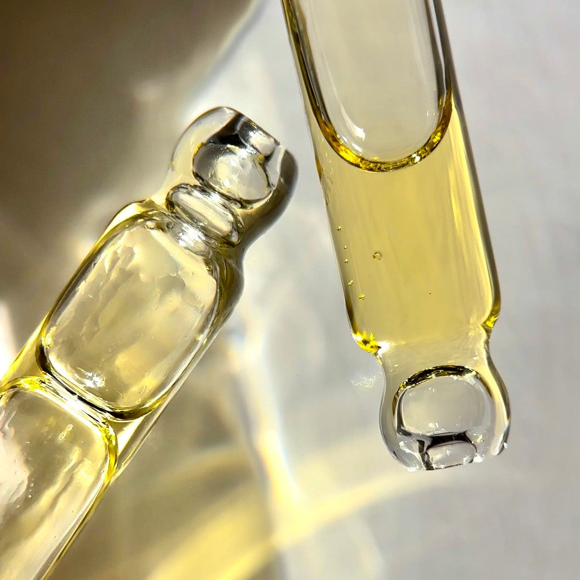 Two clear glass dropper-style vials containing pale yellow liquid, photographed close up on a reflective light surface.